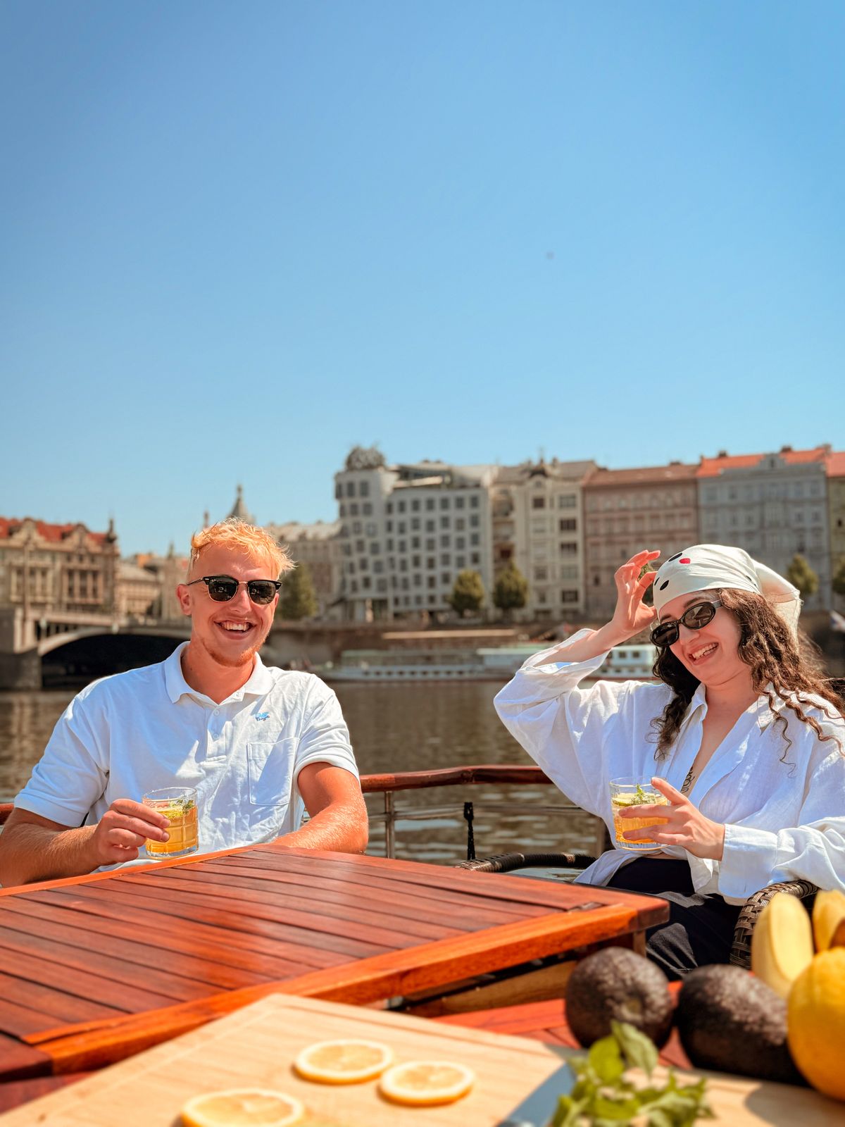 Friends with drinks at the table on a private boat in Prague, Dancing House behind